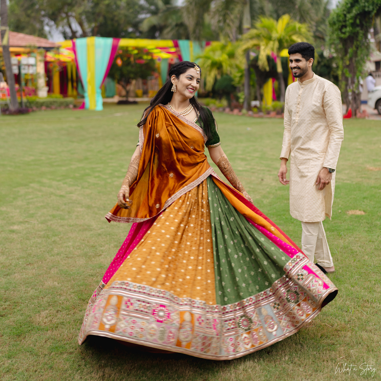 A smiling bride in a colorful mustard, green, and pink lehenga twirls on a lawn while the groom in a cream kurta looks on, with festive decorations in the background.