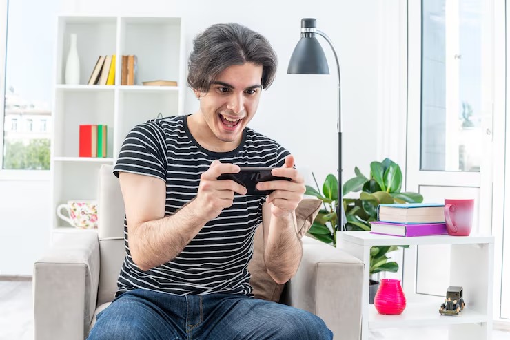 A young man sitting on a couch in a bright living room, smiling excitedly while playing a game on his smartphone, with books, a lamp, and plants in the background.